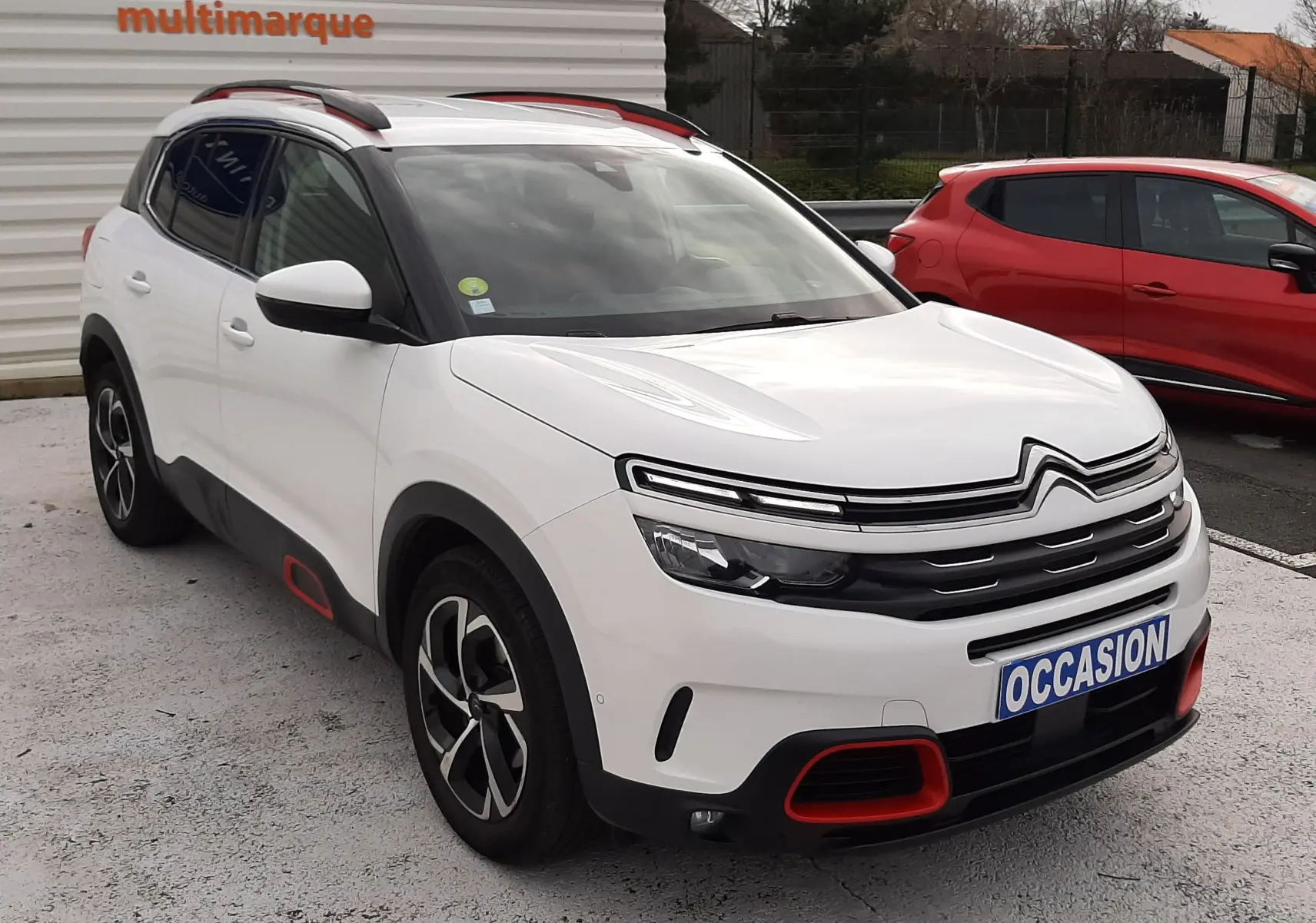 Vue 3/4 avant d'un Citroën C5 Aircross blanc avec détails rouges et jantes bicolores, stationné devant un mur blanc.