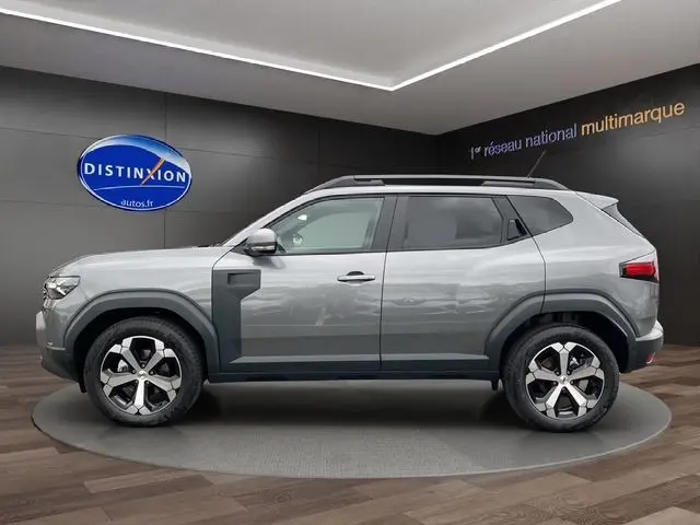 Profil côté gauche du Dacia Duster 2025 gris schiste avec jantes alliage et protections noires visibles en showroom.