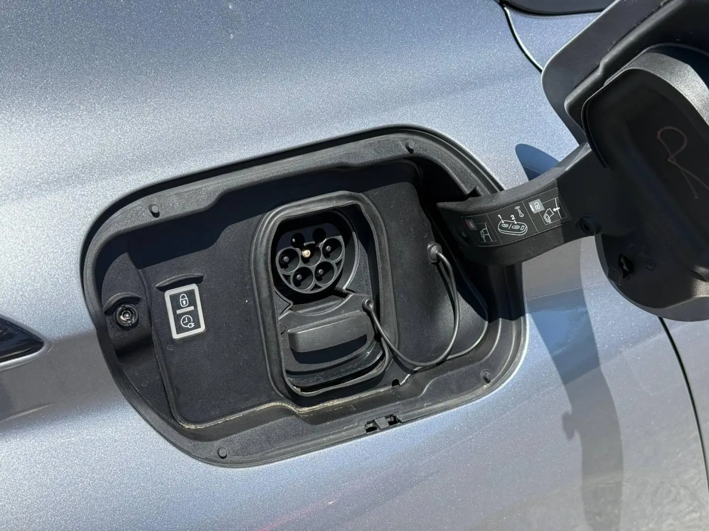 Prise de recharge électrique ouverte en gros plan sur le côté gauche d’une Peugeot 308 e-308 gris Artense.