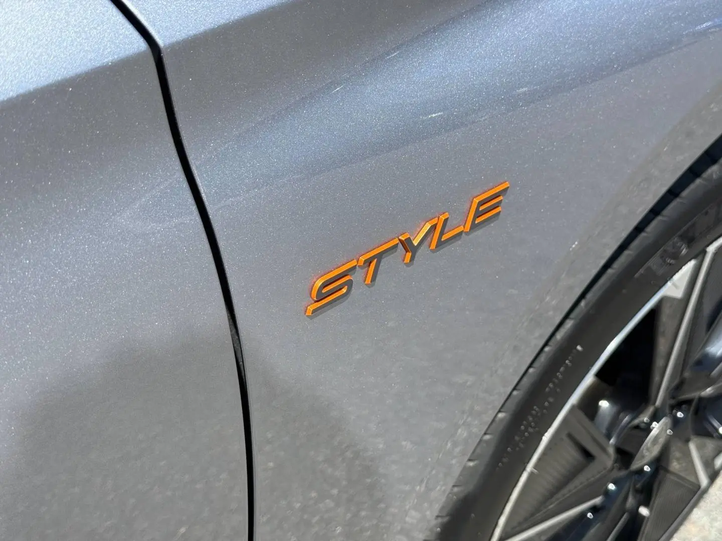 Gros plan sur l’inscription orange Style sur l’aile avant droite d’une Peugeot 308 e-308 gris Artense avec jante alu visible.