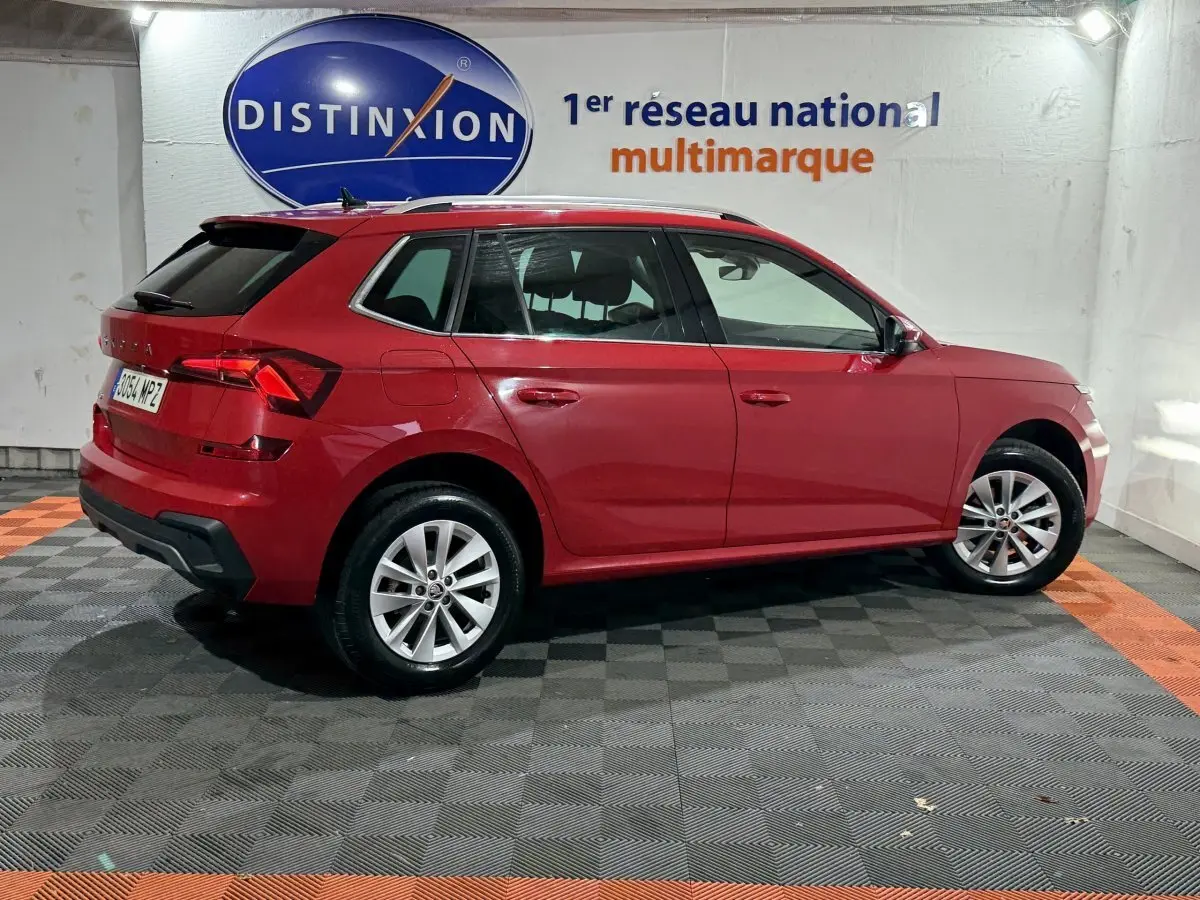 Vue latérale droite d'un Skoda Kamiq rouge 2024 avec jantes alliage et barres de toit noires en intérieur showroom.