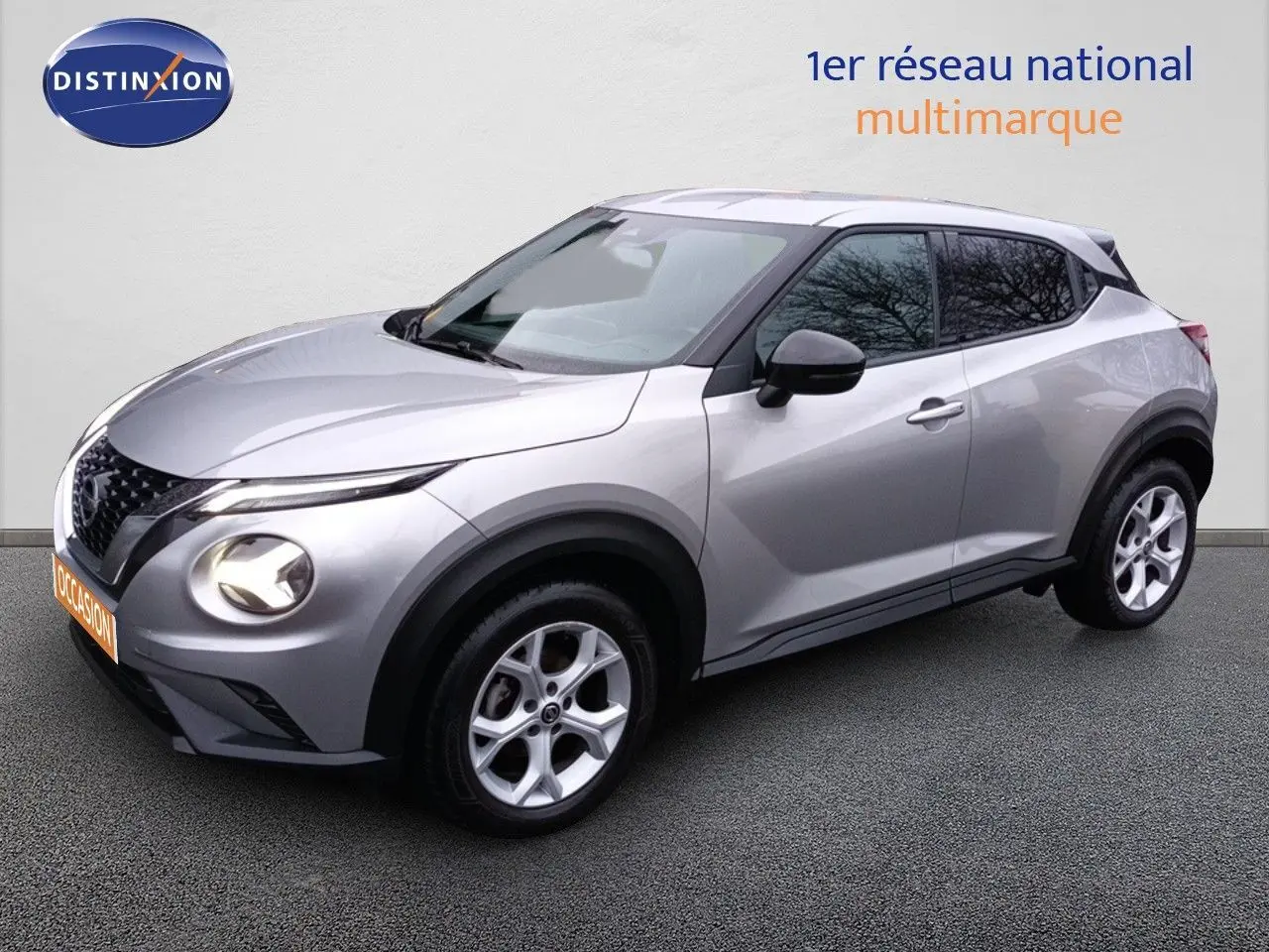 Nissan Juke gris platine métal en vue 3/4 avant droit, avec jantes alliage et phares allumés.