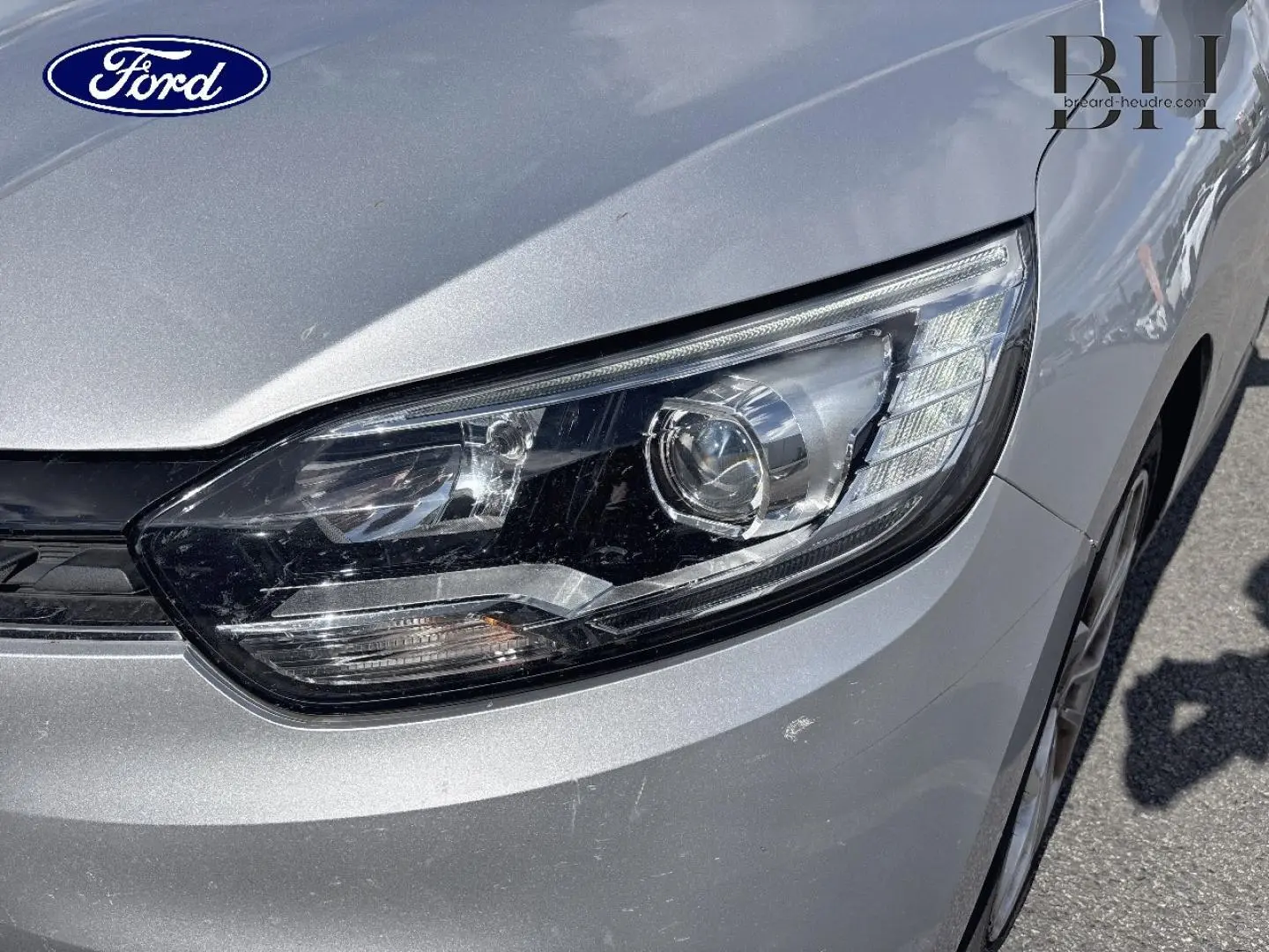 Gros plan sur le phare avant droit d'un Renault Grand Scenic gris platine, avec détails du capot et pare-chocs visibles.