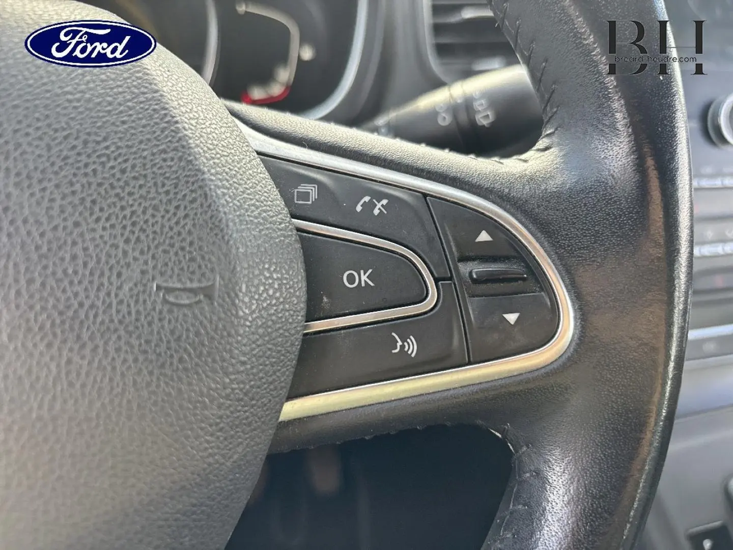 Gros plan sur le volant en cuir noir avec commandes intégrées, logo Ford visible en haut à gauche.