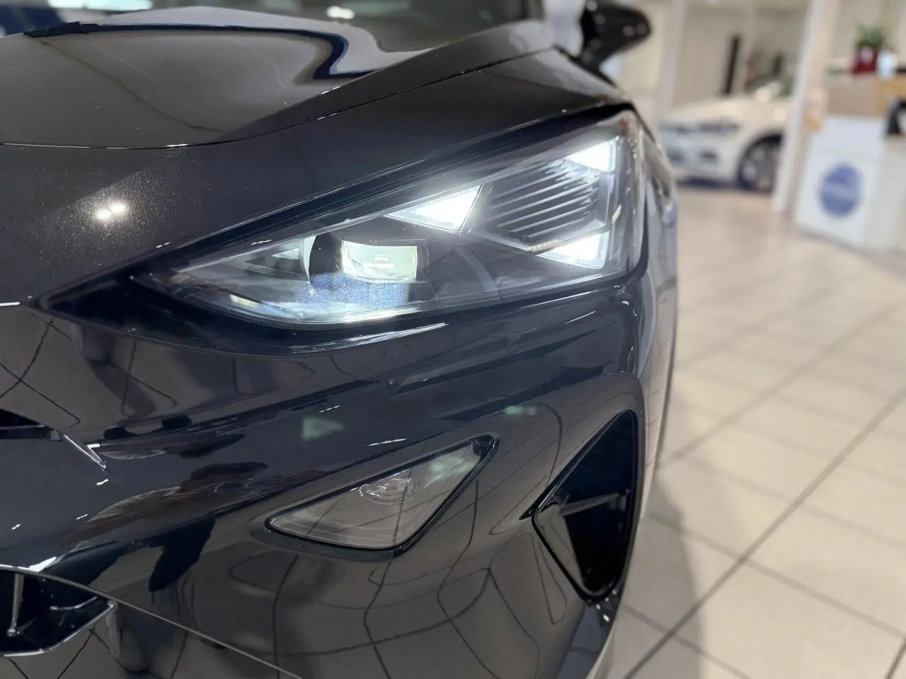 Gros plan sur le phare avant droit allumé d'une CUPRA Formentor 2025 noir Night dans un showroom.
