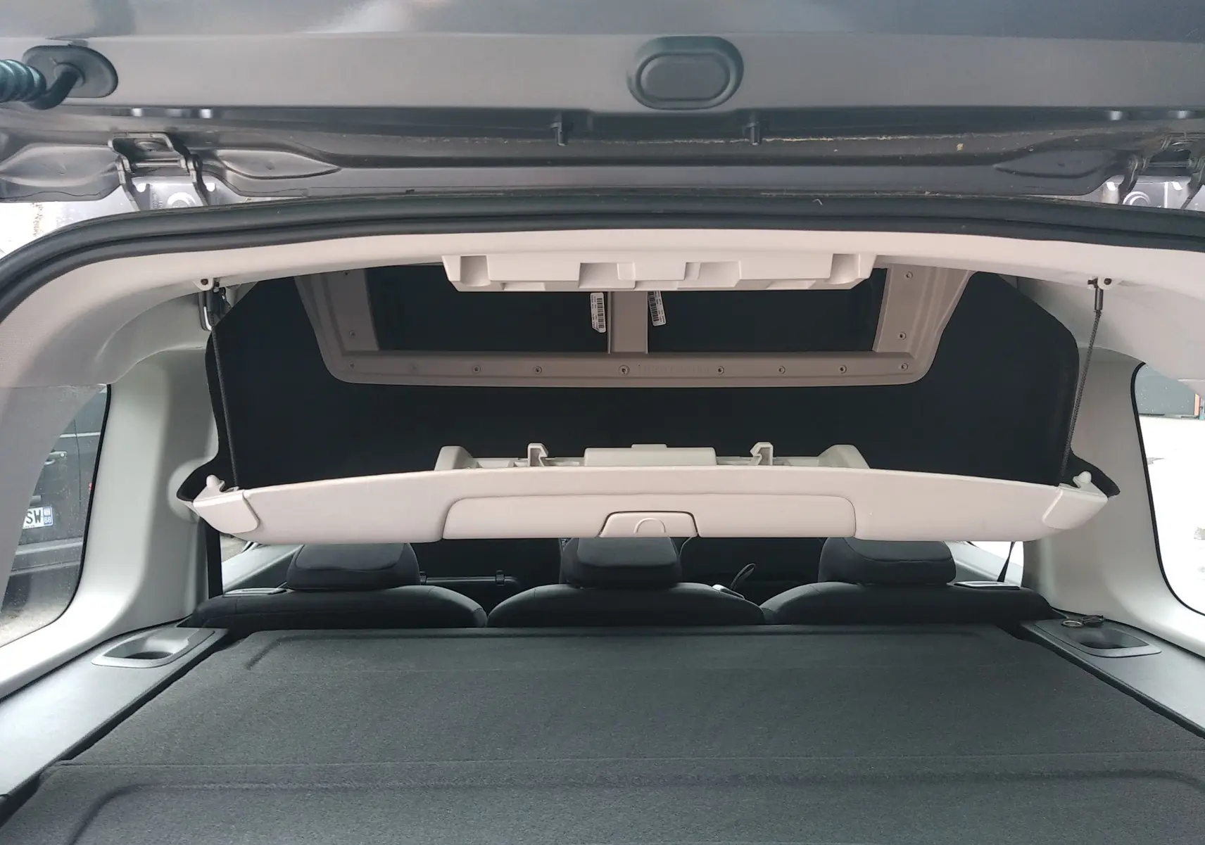 Vue intérieure arrière du coffre ouvert du Peugeot Rifter gris platinium, montrant la tablette arrière relevée et les sièges arrière noirs.