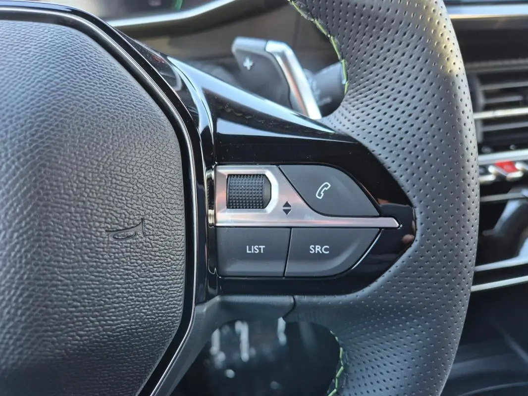 Gros plan sur la partie gauche du volant perforé noir de la Peugeot 208 Hybrid 136 e-DCS6 GT avec commandes audio intégrées.