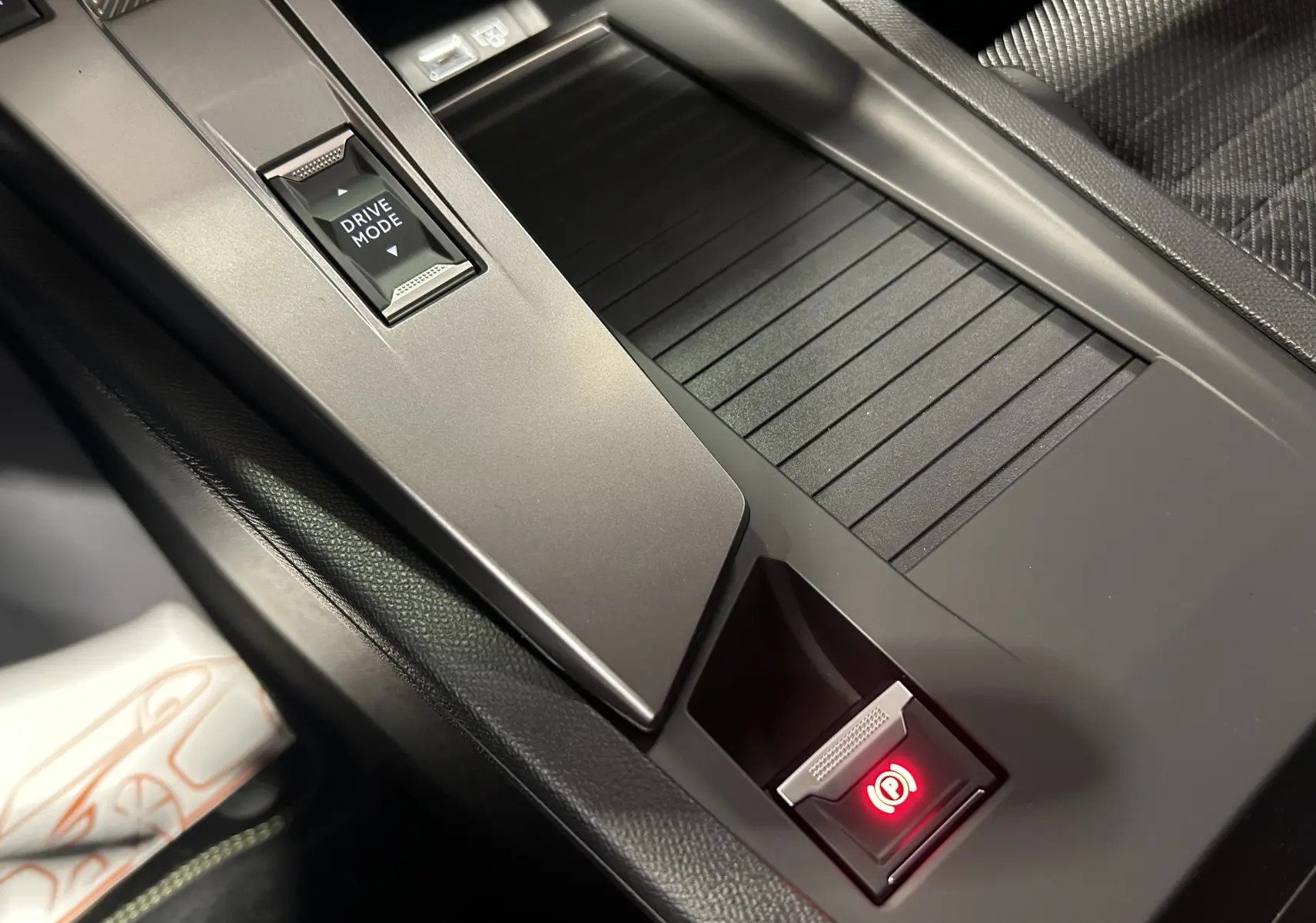 Gros plan sur la console centrale intérieure de la Peugeot 308 HYBRID 145 GT avec bouton Drive Mode et frein à main électronique rouge.