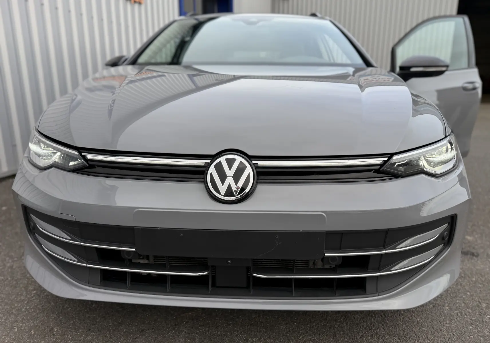Vue frontale rapprochée d’une Volkswagen Golf SW gris lunaire 2025 avec phares allumés et porte avant droite ouverte.