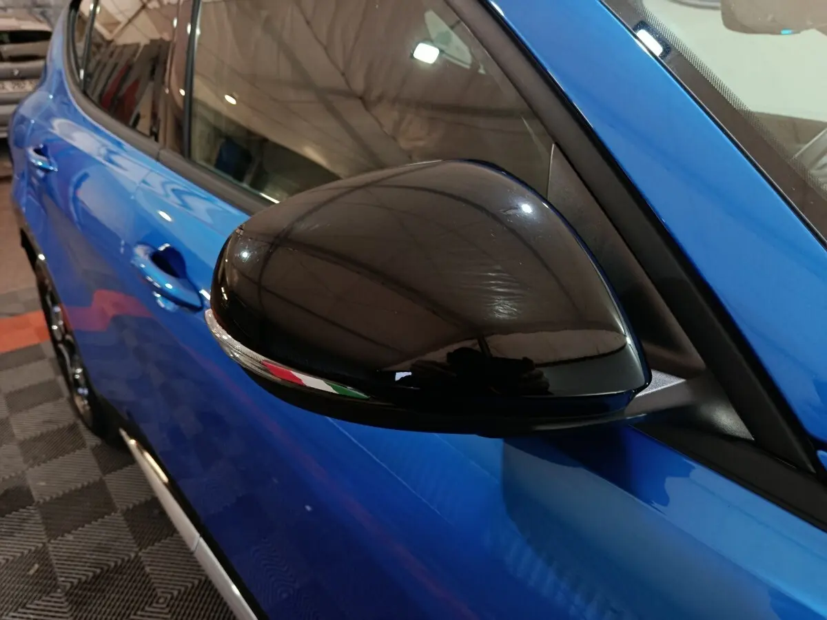 Gros plan sur le rétroviseur noir côté gauche d'une Alfa Romeo Tonale bleu 2023, avec reflet du plafond visible.