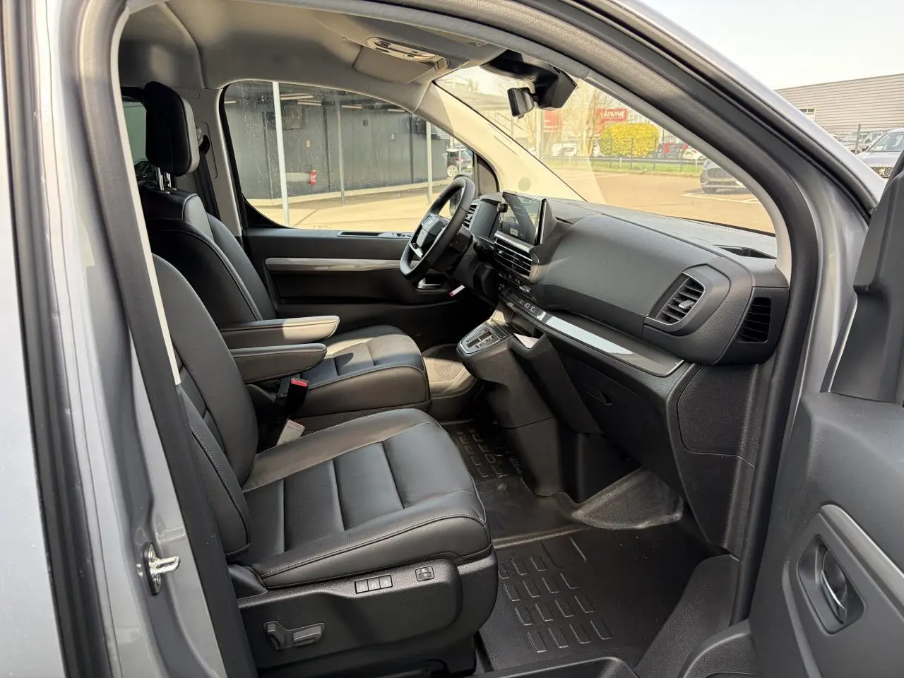 Vue intérieure côté conducteur du Citroën Jumpy gris acier, montrant sièges cuir noirs et tableau de bord moderne.
