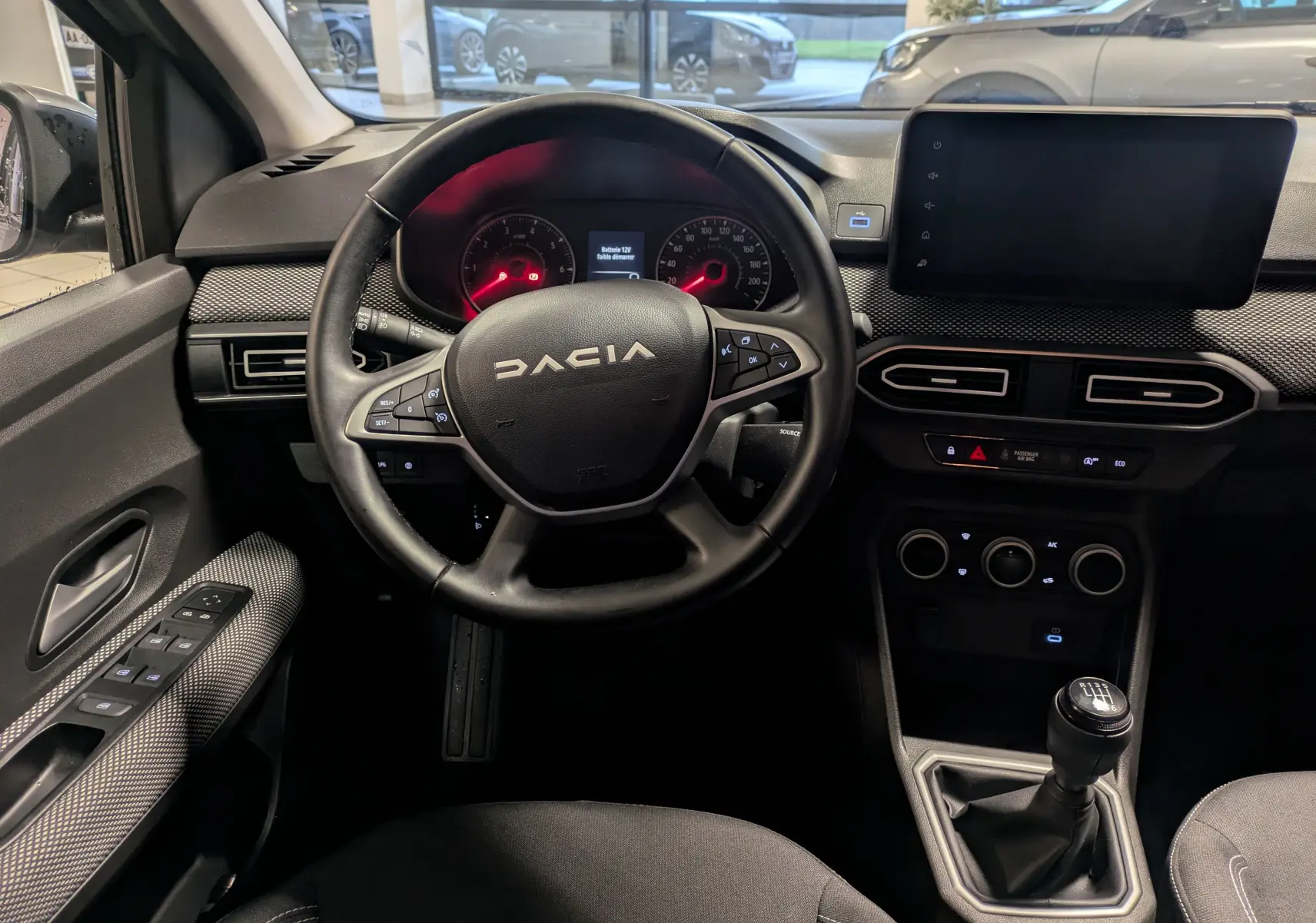 Vue intérieure centrée sur le volant DACIA et tableau de bord avec écran tactile et levier de vitesse manuel.