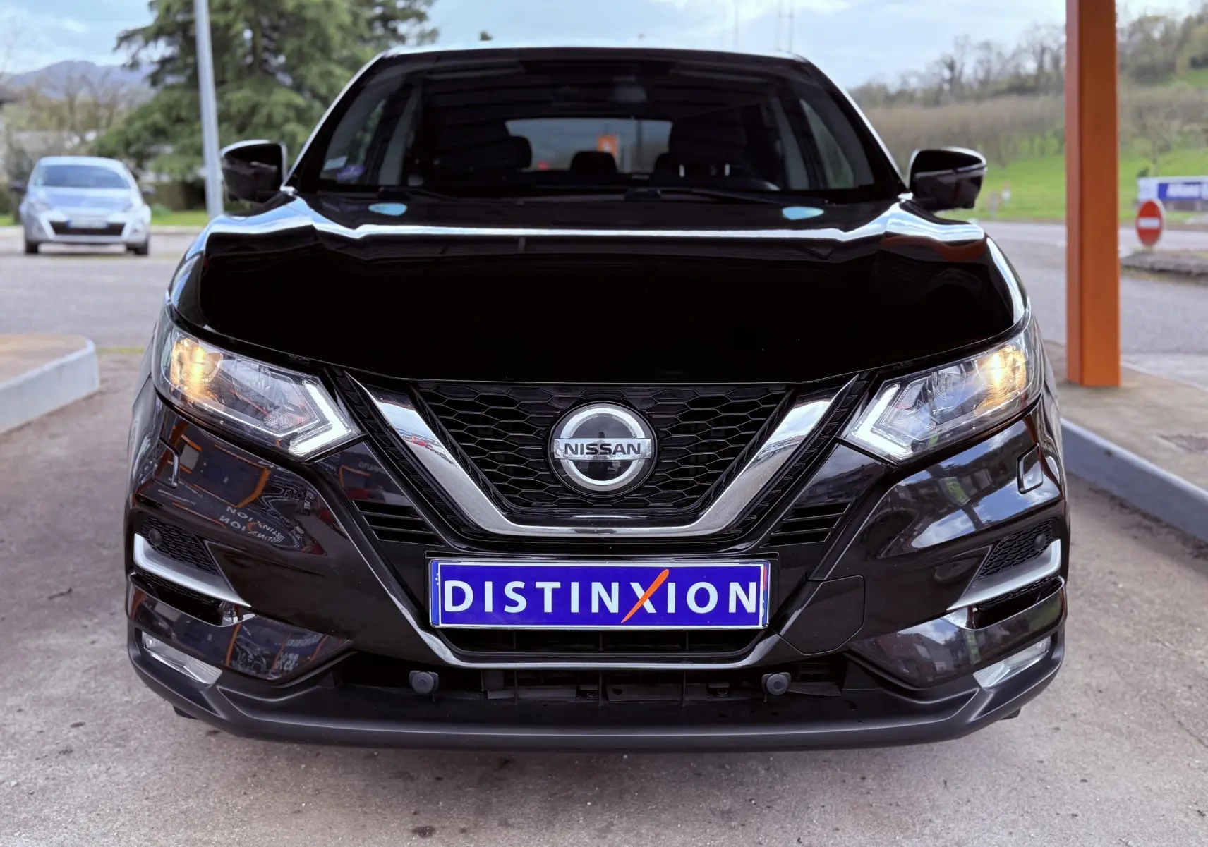 Vue frontale d'un Nissan Qashqai noir avec calandre chromée et phares allumés, plaque Distinxion visible.