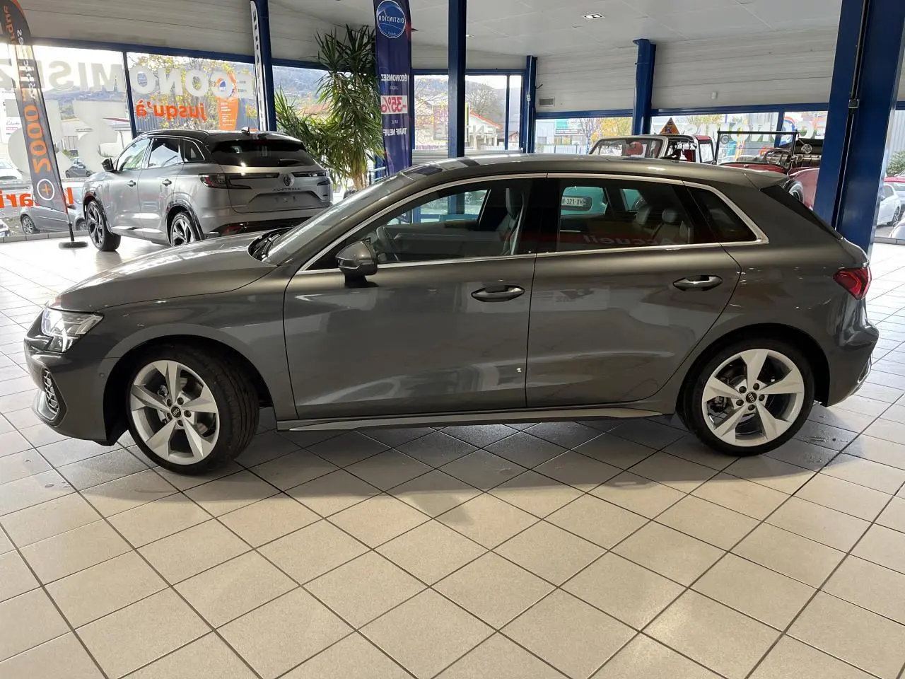 Audi A3 Sportback gris en profil droit dans un showroom, avec jantes alliage cinq branches et vitres teintées.