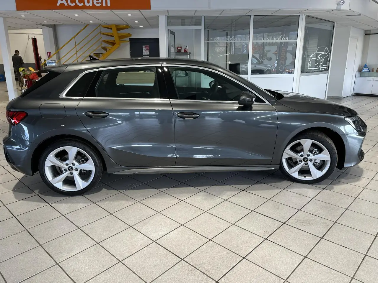 Vue de profil côté gauche d'une Audi A3 Sportback gris métallisé exposée en showroom intérieur.