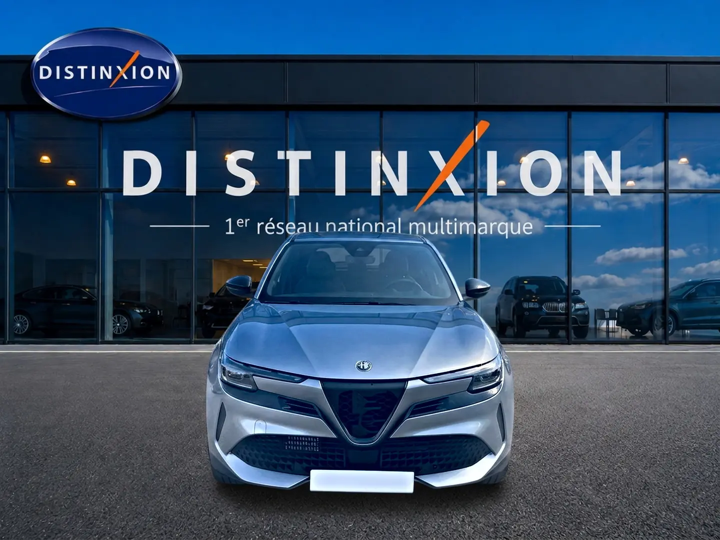 Vue frontale d'une Alfa Romeo Junior 1.2 hybride gris, avec calandre caractéristique et phares LED matrix, devant un showroom.