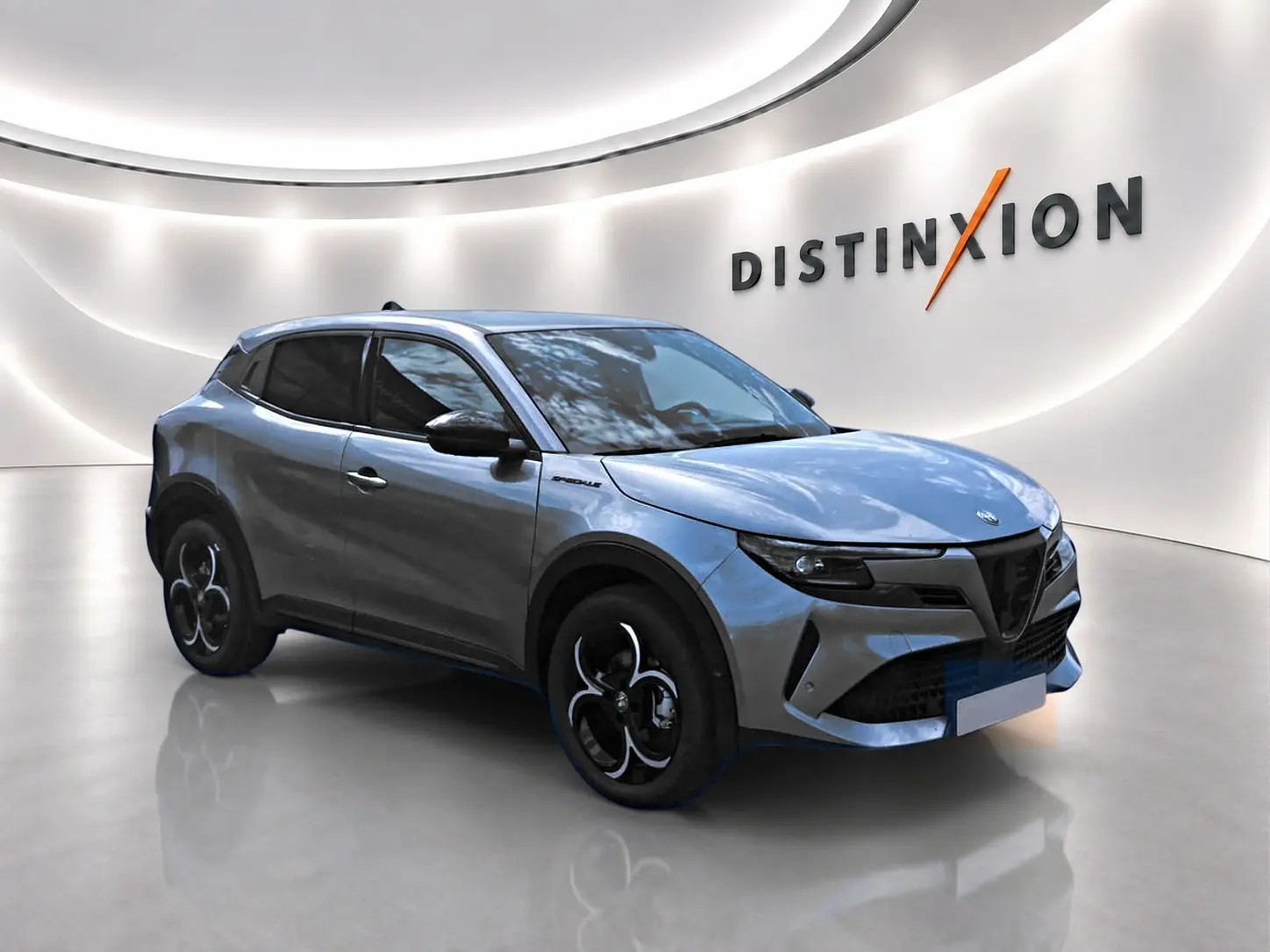 Alfa Romeo Junior gris en 3/4 avant droit, avec jantes alliage noires et vitres teintées dans un showroom lumineux.