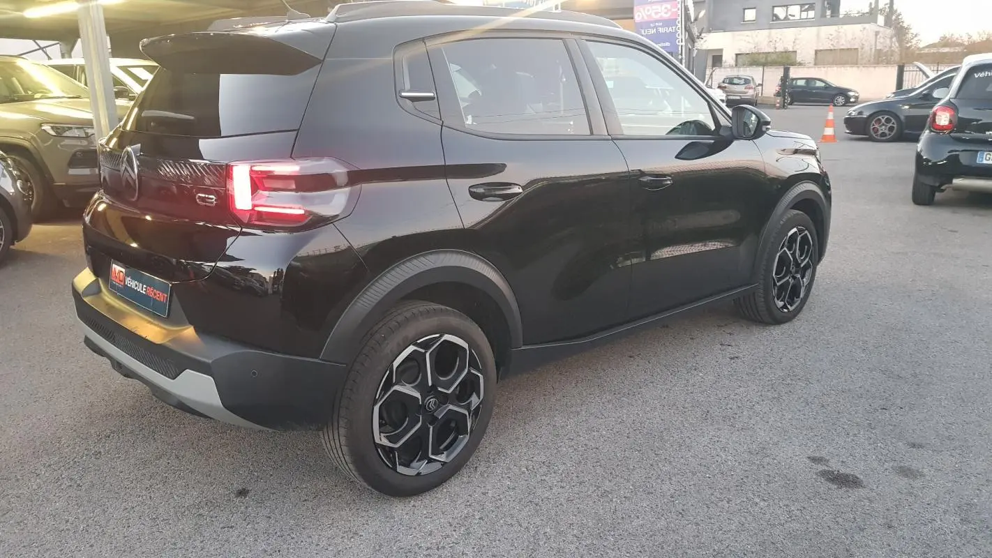 Vue 3/4 arrière droite d'une Citroën C3 noire 2025 avec jantes alliage noires et barres de toit visibles.