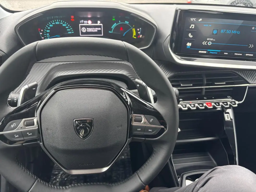 Vue rapprochée du volant et du tableau de bord numérique d'une Peugeot 208 Hybrid gris, avec écran tactile et commandes au volant.