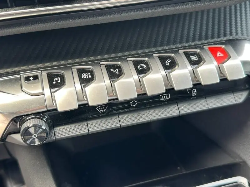 Gros plan sur la rangée de boutons métalliques du tableau de bord du Peugeot 3008 hybride bleu, avec touches fonctionnelles et alerte rouge.