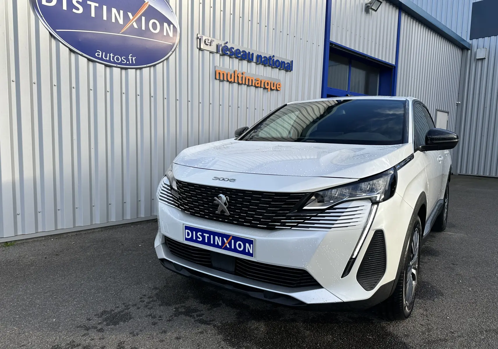 Peugeot 3008 blanc nacré en 3/4 avant droit, avec calandre noire et phares LED distinctifs, devant un bâtiment gris.