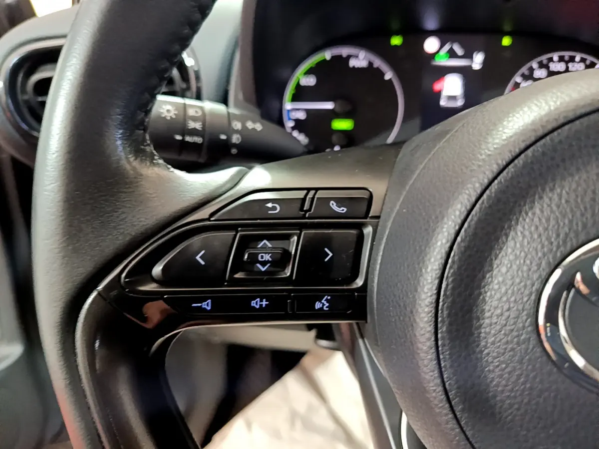 Gros plan sur les commandes du volant cuir de la Toyota Yaris hybride 2021, avec tableau de bord numérique en arrière-plan.