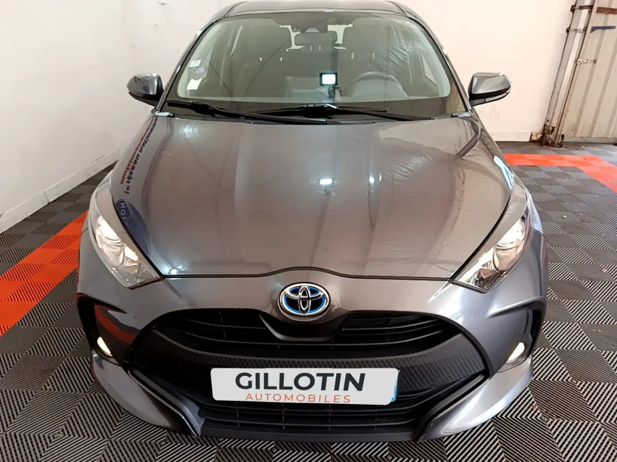 Vue frontale d'une Toyota Yaris hybride gris clair avec phares allumés dans un garage.