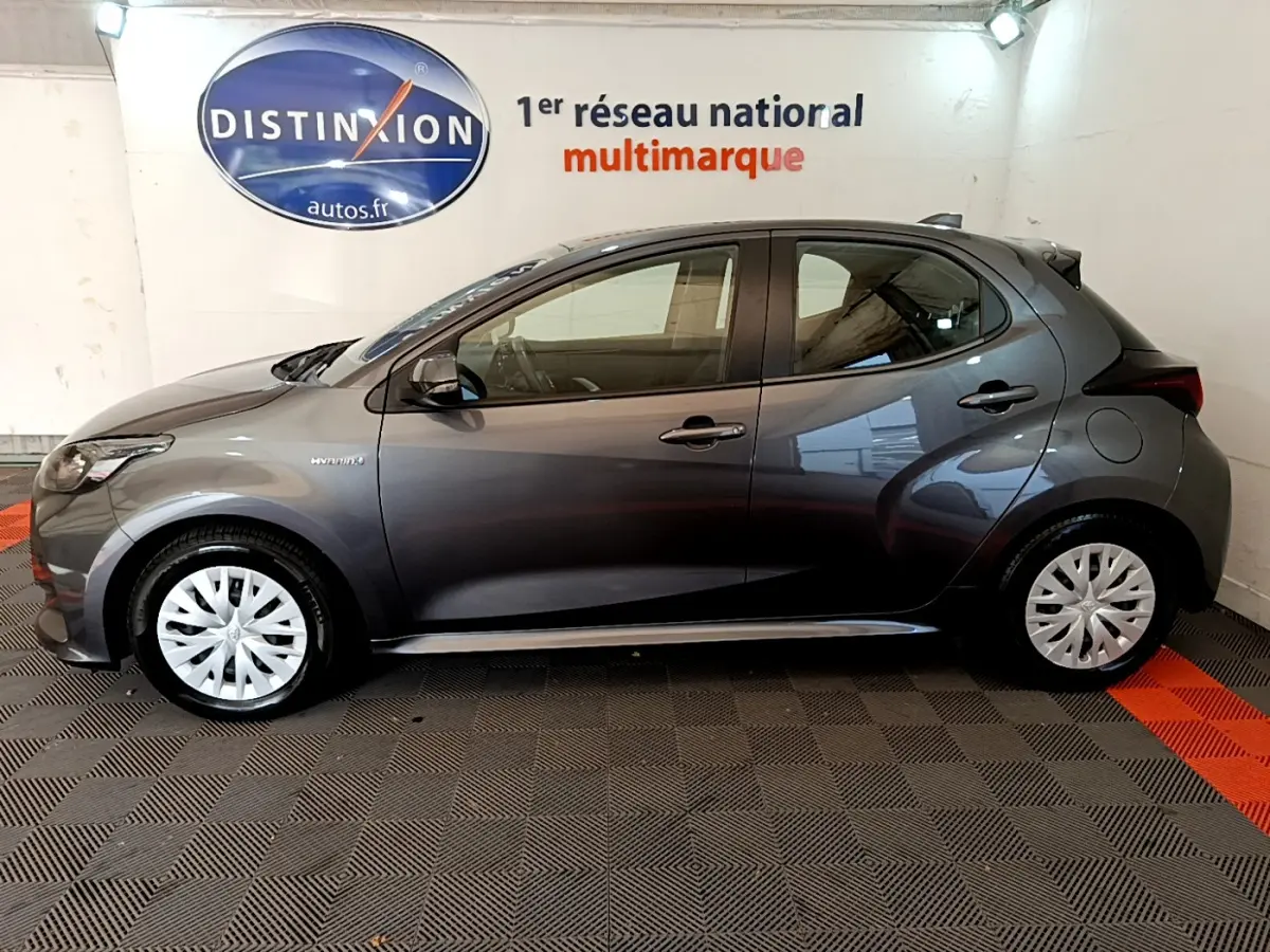 Profil côté gauche d'une Toyota Yaris hybride gris clair 2021, avec jantes à enjoliveurs blancs, en intérieur showroom.