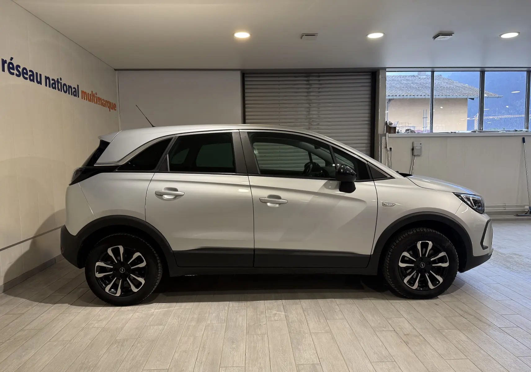 Vue latérale droite d'un Opel Crossland gris 2023 avec jantes alliage noires et toit blanc en intérieur showroom.