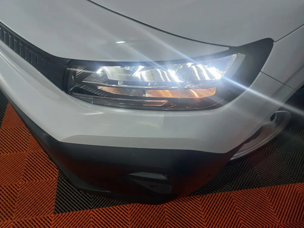 Gros plan sur le phare avant droit allumé du Citroën Berlingo XL blanc, avec pare-chocs noir visible.