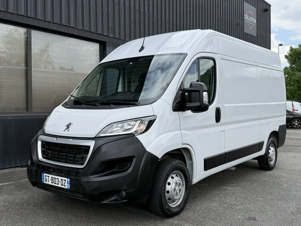 Vue 3/4 avant droit d’un Peugeot Boxer Fourgon blanc avec pare-chocs noir et rétroviseurs noirs, stationné devant un bâtiment.