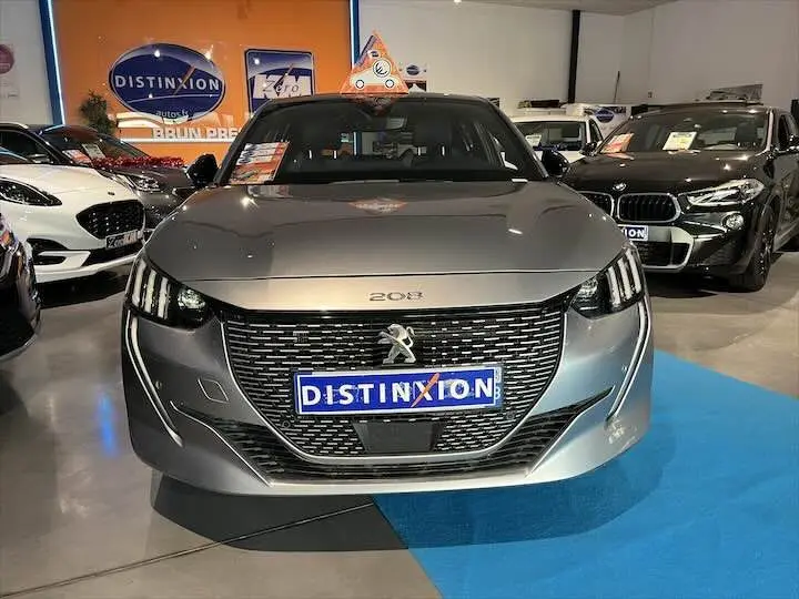 Vue frontale d'une Peugeot 208 E-208 GT gris Artense exposée en showroom avec calandre noire et phares LED distinctifs.