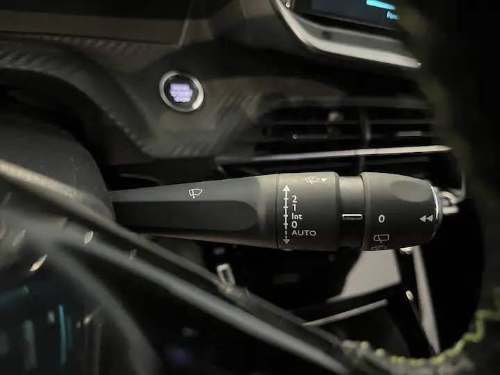 Gros plan sur la commande des essuie-glaces à gauche du volant dans l'habitacle sombre de la Peugeot e-208 gris Artense.
