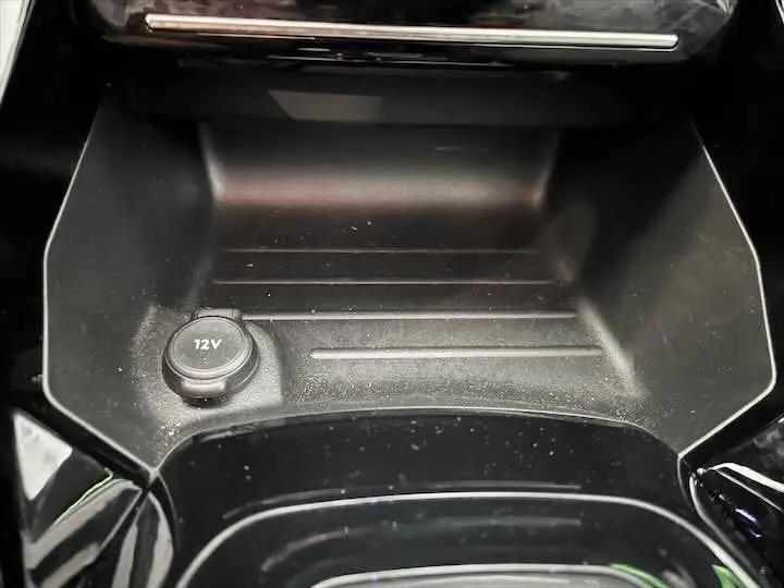 Prise 12V et rangement central noir mat dans l'habitacle de la Peugeot e-208 GT gris Artense, vue rapprochée.