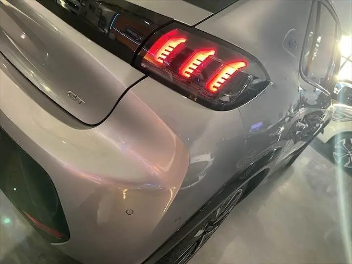 Vue 3/4 arrière droite d'une Peugeot 208 E-208 GT gris Artense avec feux arrière LED allumés et logo GT visible.