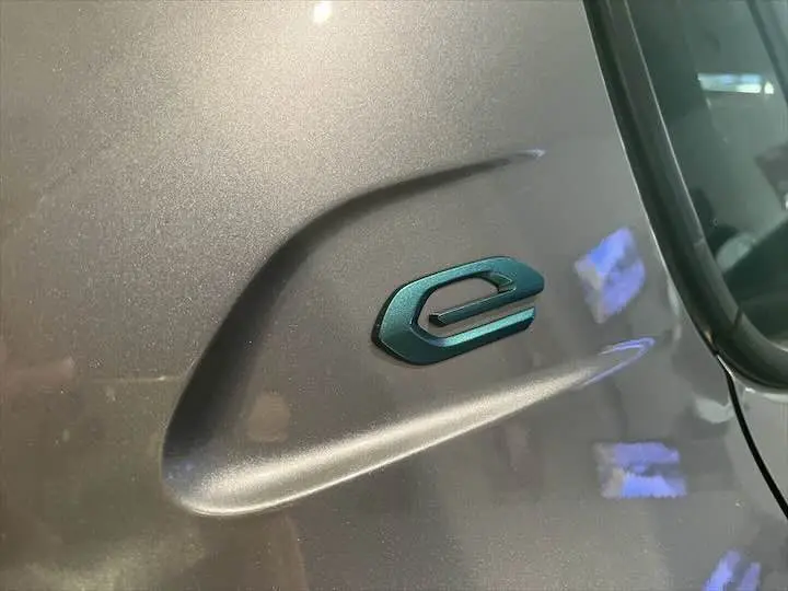Gros plan sur l'emblème "e" bleu sur la carrosserie gris Artense de la Peugeot 208 électrique côté avant droit.
