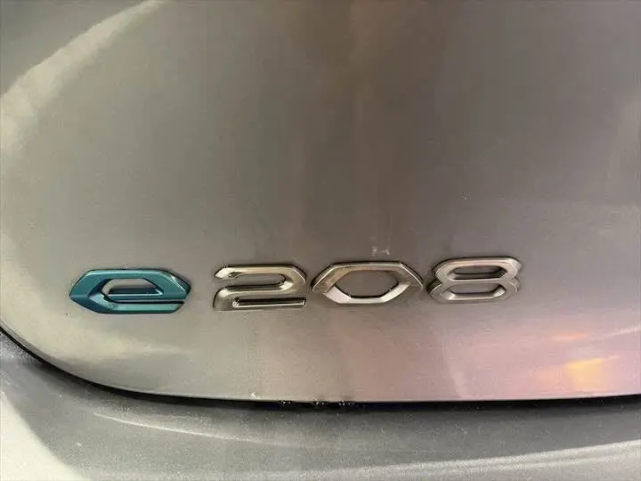 Gros plan sur l'emblème e-208 bleu et argent sur la malle arrière gris artense d'une Peugeot électrique.