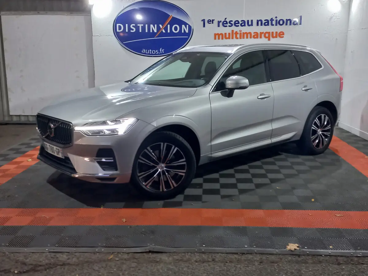 Volvo XC60 B4 gris clair vu de 3/4 avant droit, avec jantes alliage et phares LED allumés en intérieur showroom.