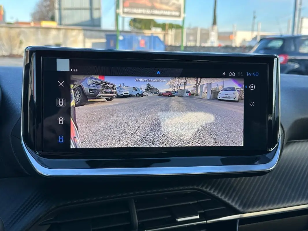 Écran tactile intérieur montrant la caméra de recul d'une Peugeot 208 blanche, vue arrière sur parking.