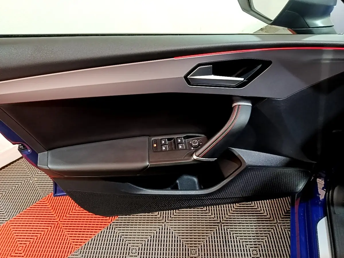 Vue intérieure côté gauche de la porte avant d'une SEAT Leon bleu, avec poignée cuir et surpiqûres rouges.