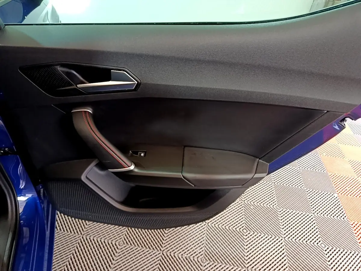 Vue rapprochée de la porte avant gauche bleu de la SEAT LEON 1.5 eTSI avec poignée noire et surpiqûres rouges.
