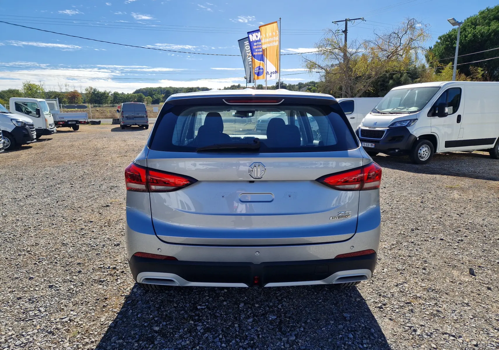 Vue arrière d'un MG Motor 1.5 L Hybrid+ Cosmic Silver garé sur un sol gravillonné, feux arrière LED visibles.