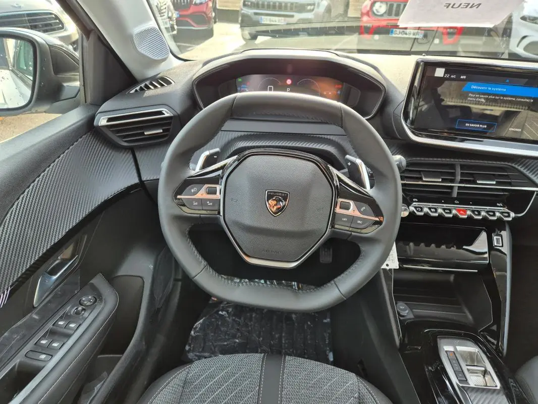 Vue intérieure centrée sur le volant et le tableau de bord de la Peugeot 208 Hybrid blanc, avec écran tactile et finitions carbone.