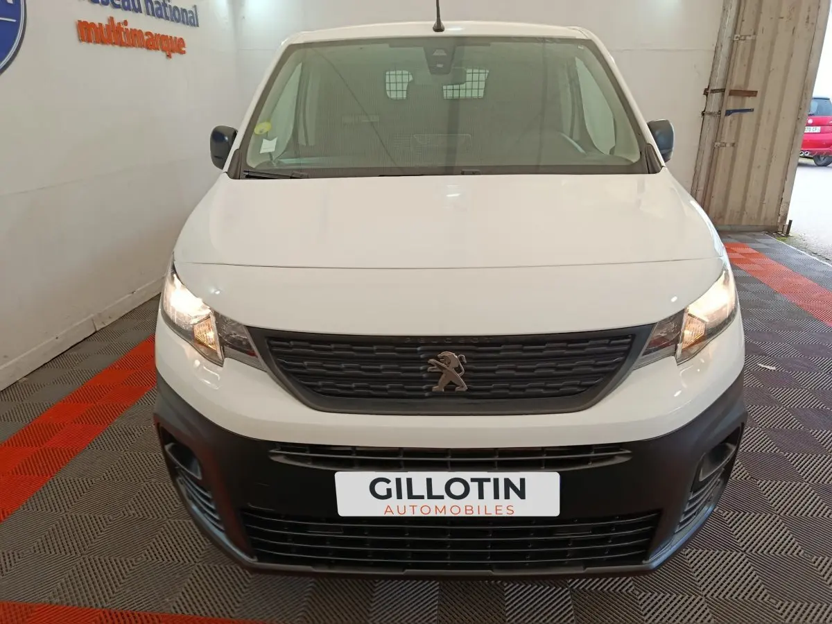 Vue avant d'un Peugeot Partner blanc avec calandre noire et phares allumés dans un garage.