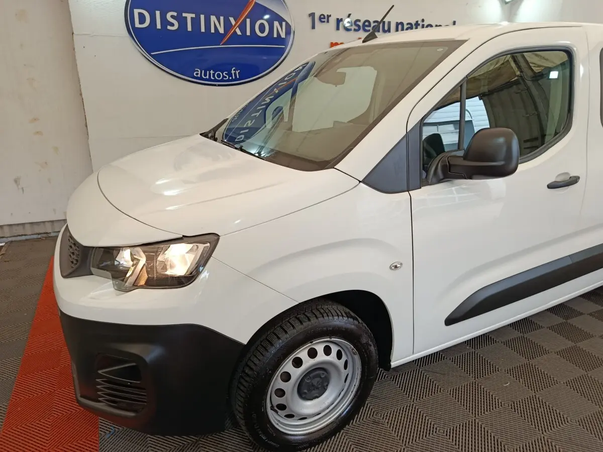 Vue 3/4 avant gauche d’un Peugeot Partner blanc avec pare-chocs noir et jante acier simple en intérieur showroom.