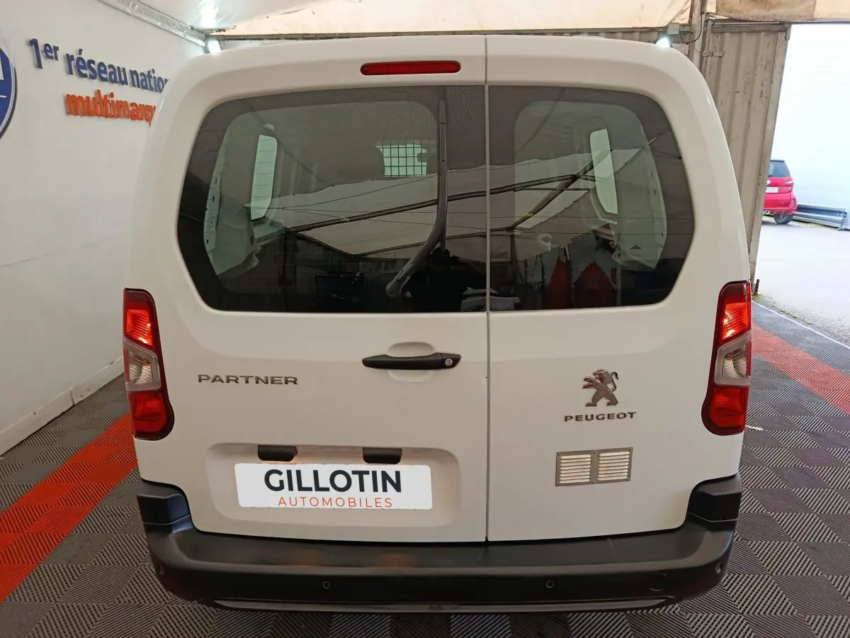 Vue arrière d’un Peugeot Partner blanc 2021 avec portes battantes et logo visible, stationné en intérieur.