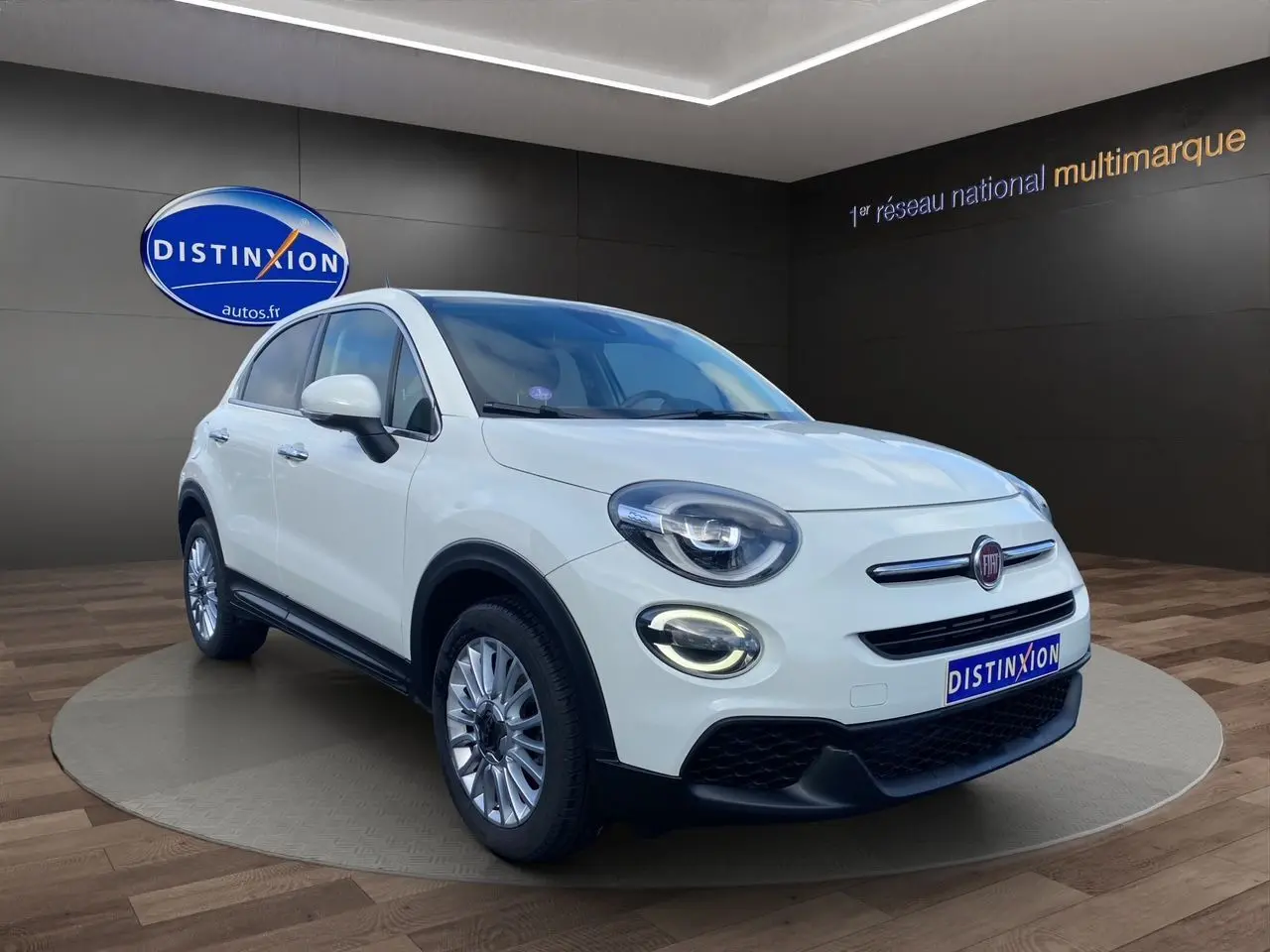 Fiat 500X blanc en 3/4 avant droit, avec phares LED allumés et jantes alliage 17 pouces dans un showroom.