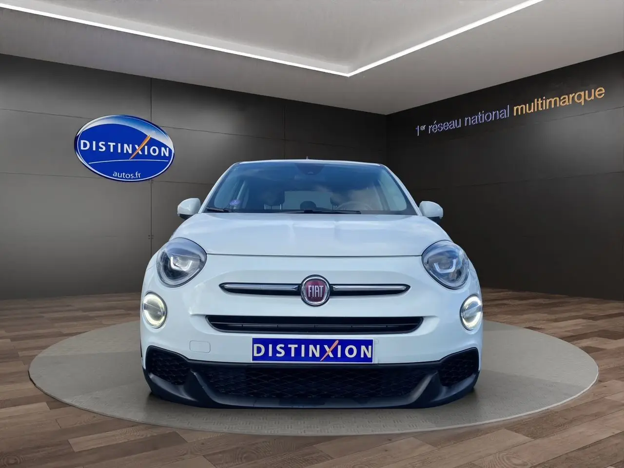 Vue frontale d'une Fiat 500X blanche avec phares allumés et logo FIAT bien visible sur un fond intérieur sombre.
