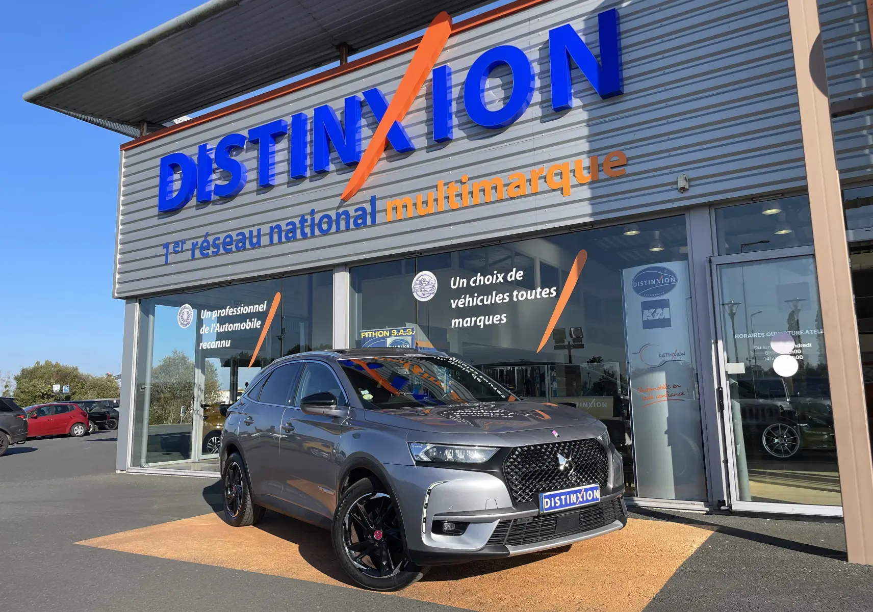 DS7 gris Artens en 3/4 avant droit, avec calandre noire distinctive et jantes noires brillantes, devant un showroom Distinxion.