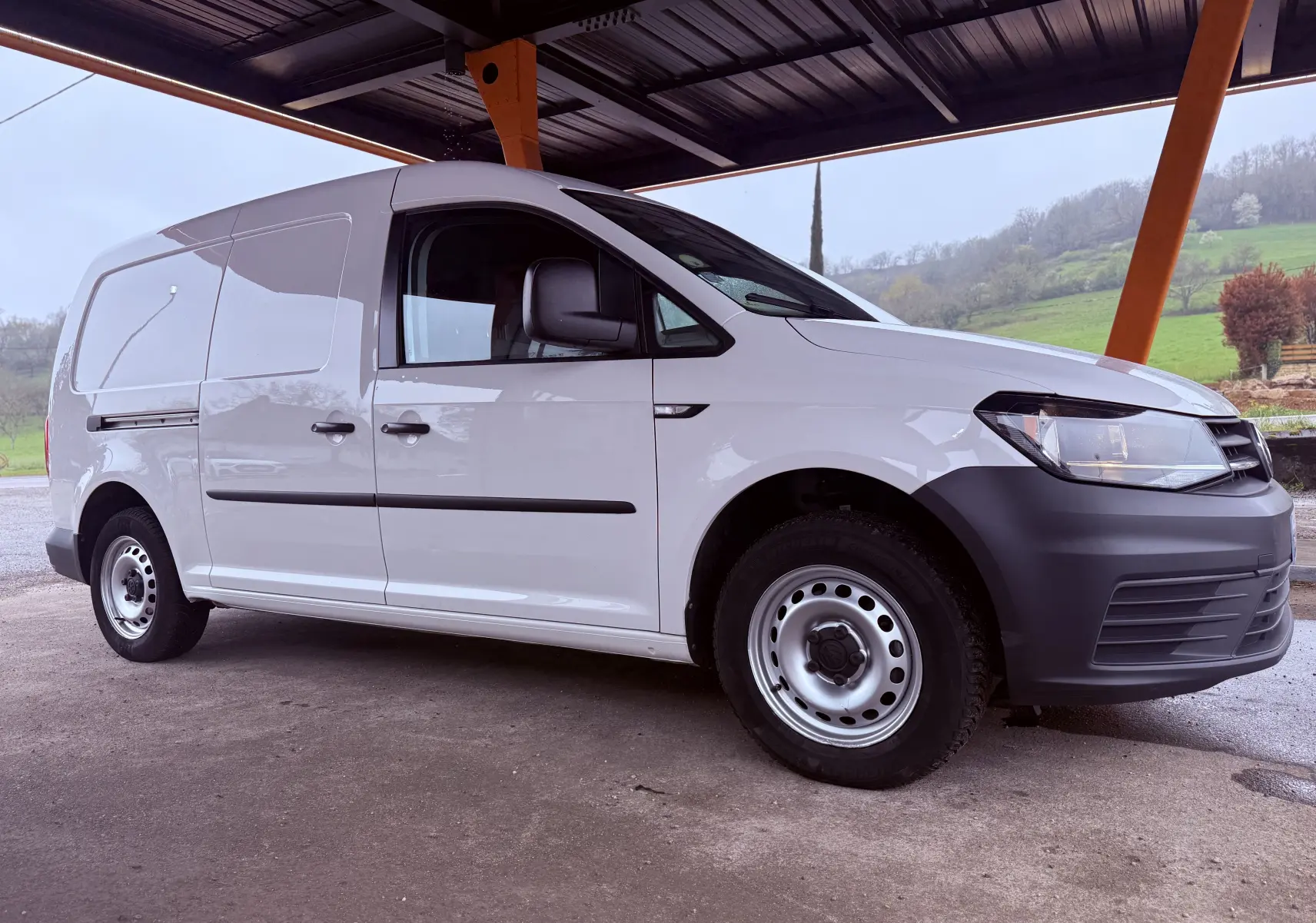 Vue 3/4 avant droit d’un Volkswagen Caddy Maxi Long blanc avec pare-chocs noirs sous un abri en milieu rural.